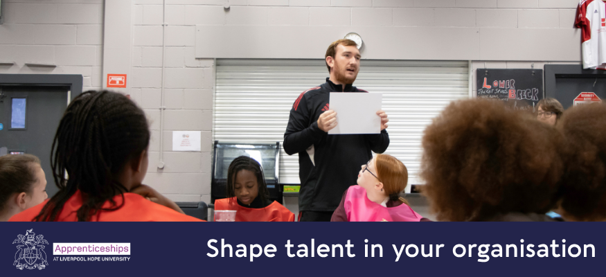A male youth worker stands in the middle of a youth centre in front of a group of children who are crafting and colouring. He is holding up a piece of artwork to display to the room.